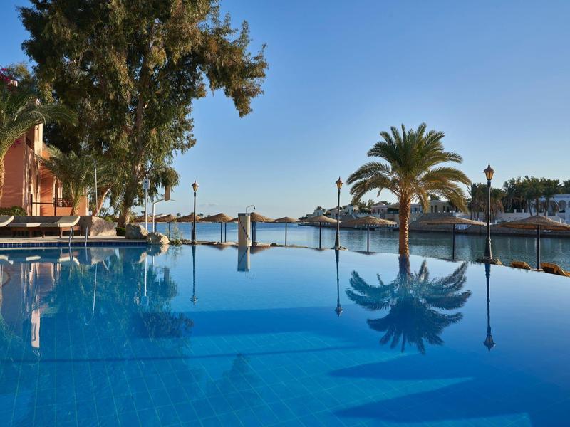 Piscine calme avec chaises et palmiers sous un ciel bleu clair près de l'eau et des bâtiments.