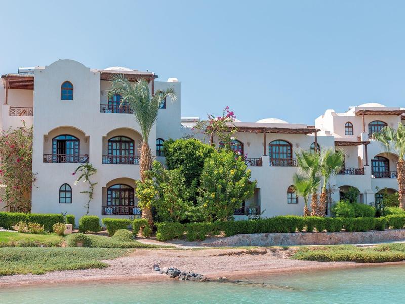 Hôtel blanc près d'un lagon turquoise sous un ciel bleu clair avec des palmiers et de la verdure.