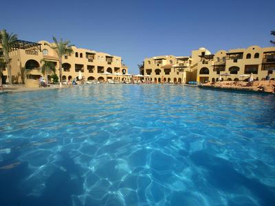 Grande piscina all'aperto davanti a un edificio alberghiero su più piani sotto un cielo azzurro.