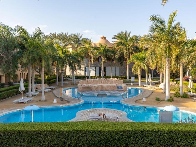 Großer blauer Pool mit Wasserfall, umgeben von Palmen und Gebäude im Hintergrund.