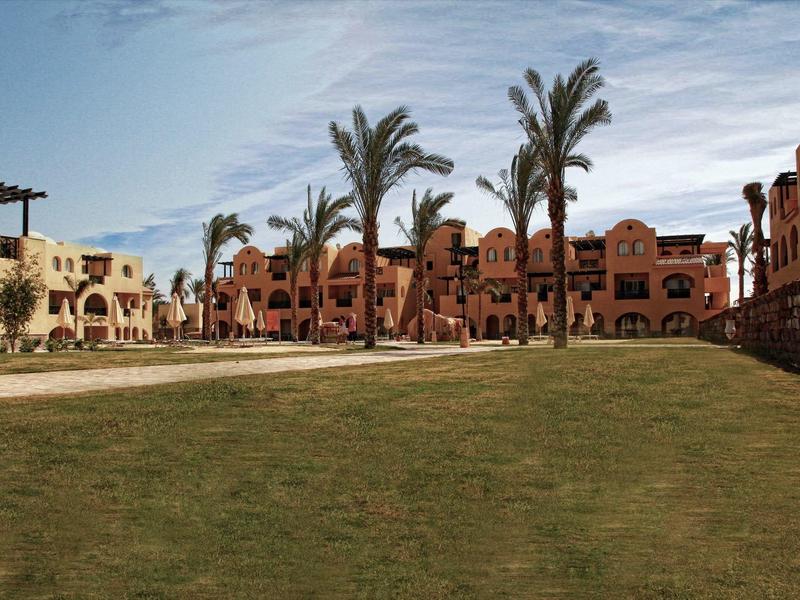 Große Rasenfläche zwischen mehrstöckigen Gebäuden mit Palmen unter blauem Himmel.