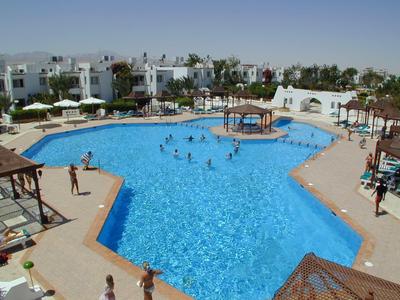 Großer Pool mit klarem Wasser, umgeben von weißen Gebäuden und Sonnenschirmen, Menschen schwimmen.