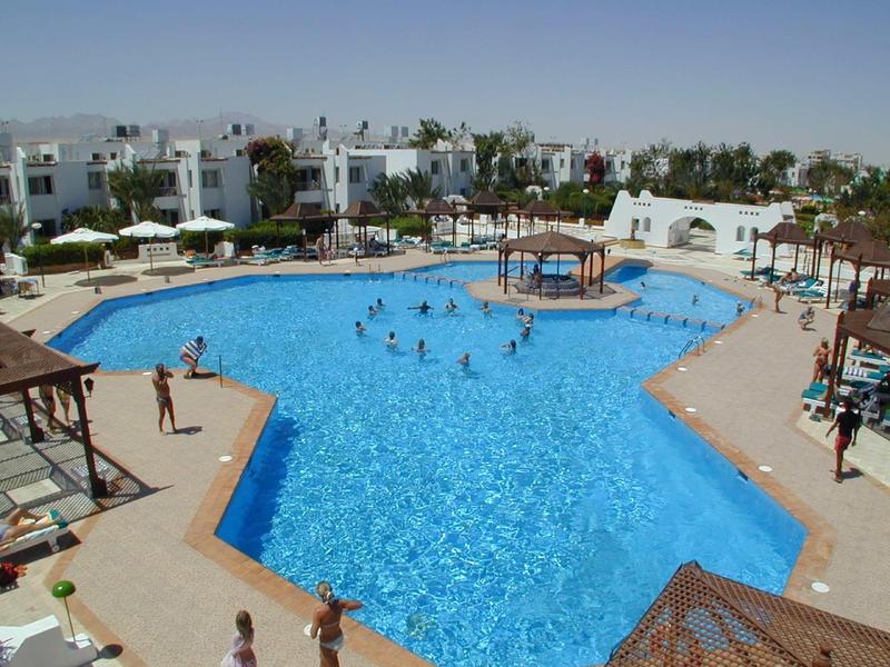 Grande piscine d'hôtel avec de nombreux nageurs et des bâtiments blancs environnants et des palmiers.