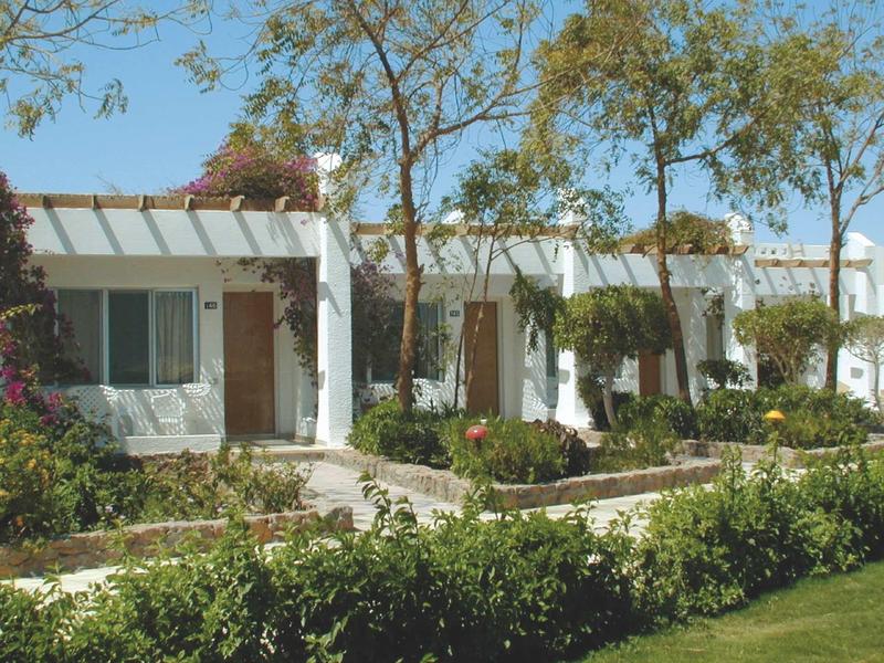Bungalows blancs de vacances avec portes et fenêtres vertes entourés d'arbres et de jardins bien entretenus.