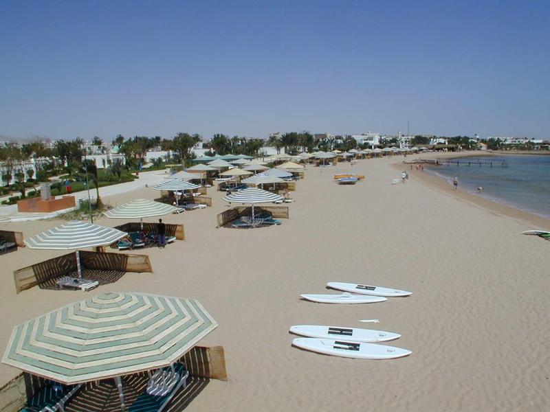 Large plage de sable avec parasols et planches de surf, mer calme et ciel bleu clair.