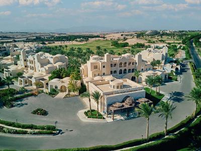 Resort with beige buildings, palm trees, and a road surrounded by green fields under a cloudy sky.