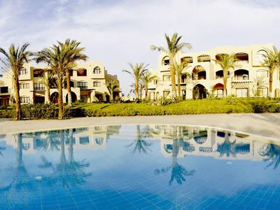 A hotel building with palm trees reflected in the blue swimming pool under a clear sky.