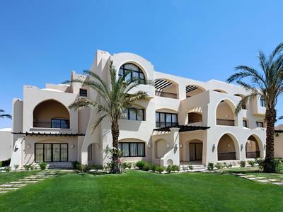 White hotel building with arches and palm trees under blue sky on green lawn.