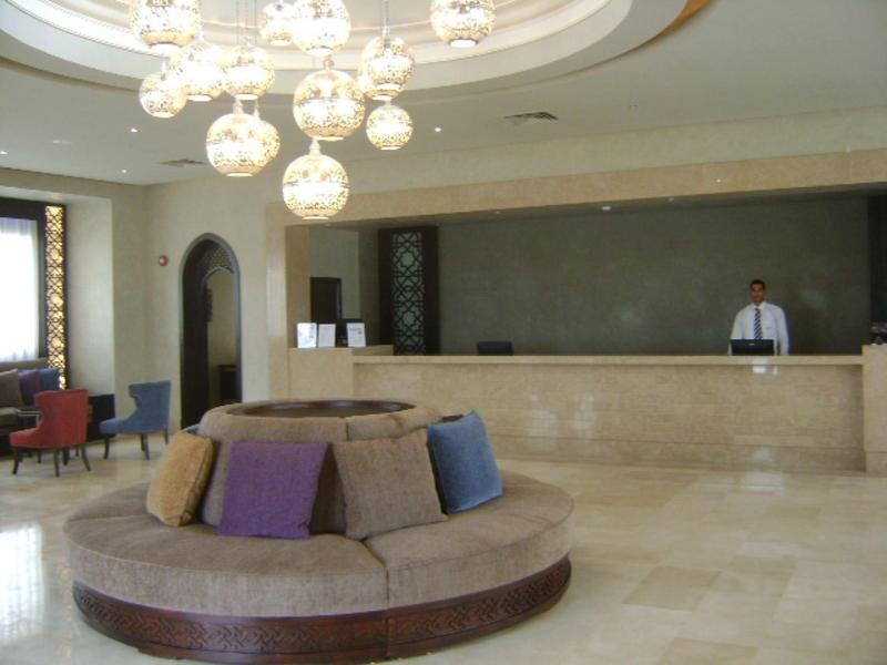 Hotel lobby with a circular beige sofa, colorful pillows, chandelier, and a reception desk with a staff member.