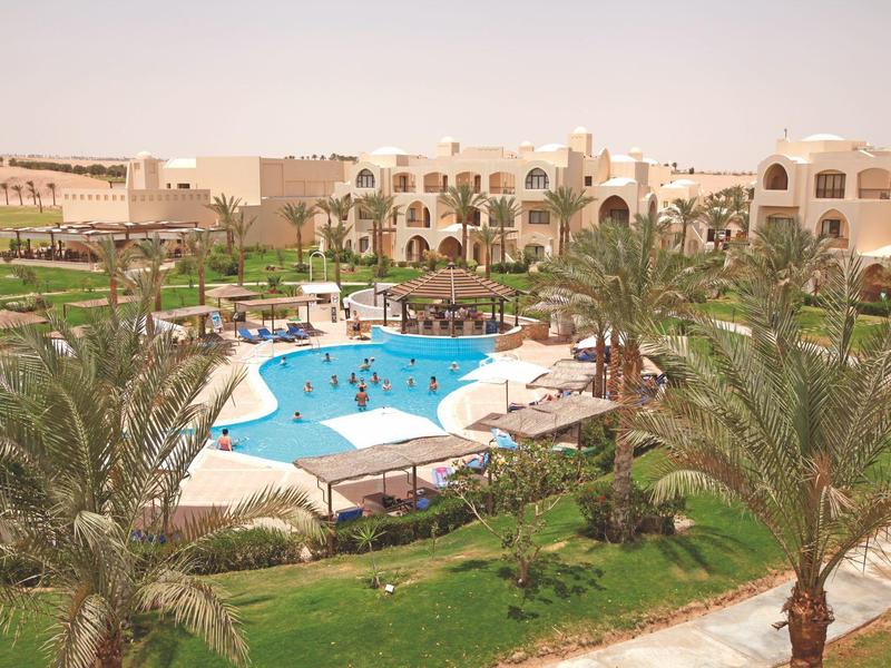 Resort with palm trees, blue swimming pool, and beige multi-story buildings under a clear sky.