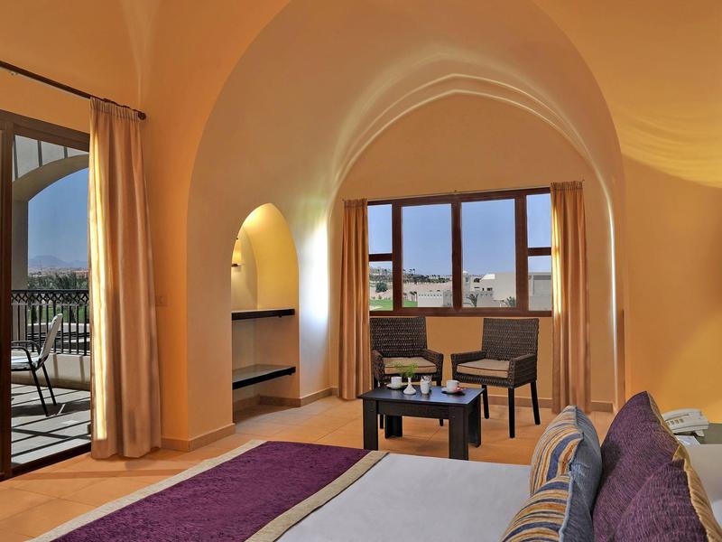 Cozy hotel room with arched ceiling, bed, sitting area, and balcony view at sunset.