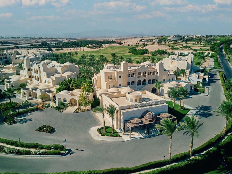 Resort with beige buildings, palm trees, and a road surrounded by green fields under a cloudy sky.