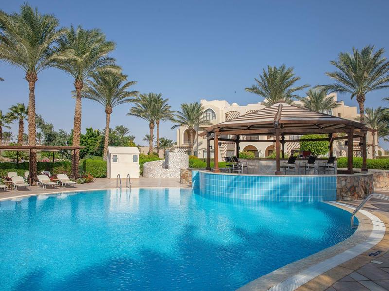 Outdoor pool with bar surrounded by palm trees and lounge chairs under a clear sky.