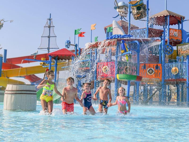Children playing in a colorful water park with slides and splash structures under a clear sky.