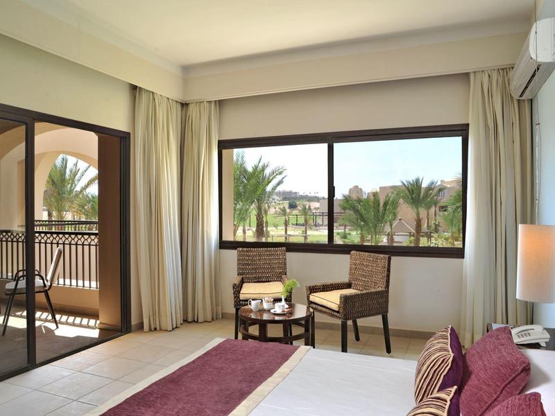 Hotel room with large window showing palm trees, balcony with chairs, and cozy bed with red accents.