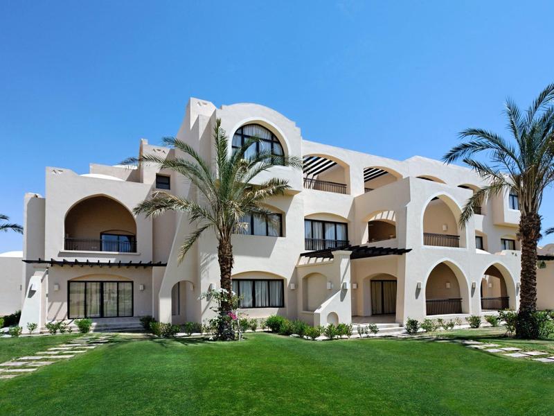 White Mediterranean hotel building with arches, palm trees, and green lawn under clear blue sky.