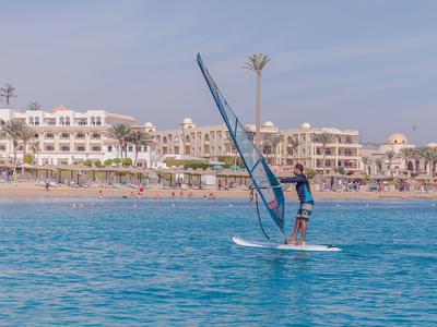 Person surft auf einem weißen Board mit blauem Segel auf klarem, blauem Wasser nahe Strand mit Hotels.