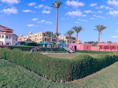 Parco giochi verde con siepi e palme davanti a edifici sotto un cielo azzurro.