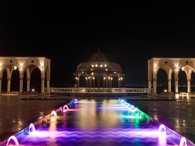 Piscina illuminata con fontane colorate e un grande edificio di notte.