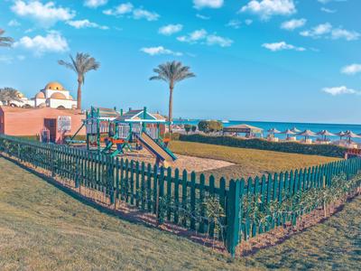 Un'area giochi recintata con un'altalena davanti a un hotel sul mare in una giornata di sole.