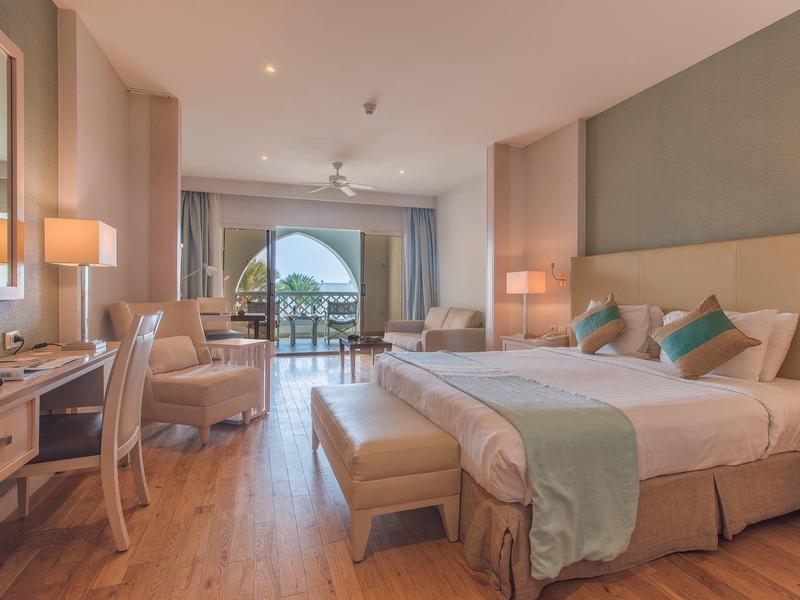 Prostorný hotelový pokoj s manželskou postelí, psacím stolem, posezením a balkonem s výhledem na moře.