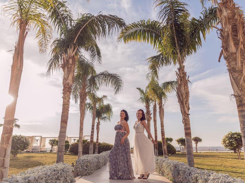 Dvě ženy v dlouhých šatech stojí mezi palmami na slunečné cestě.