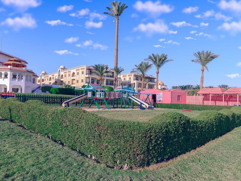 Zelený park s živým plotem, hřištěm a palmami před vícepodlažními budovami pod modrou oblohou.