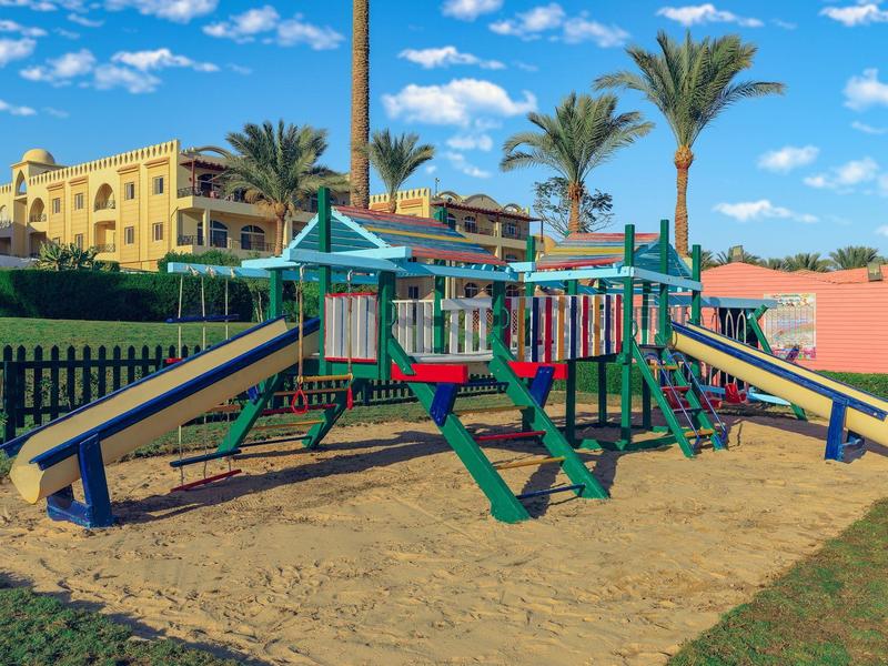 Barevné dětské hřiště s klouzačkami a lezeckými sítěmi před hotelovou zahradou a modrou oblohou.