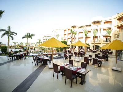 Outdoor hotel dining area with tables, chairs, yellow umbrellas, palm trees, and a multi-story building.