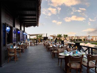 Otevřená restaurace s dřevěnou podlahou a výhledem na palmy při západu slunce.