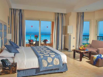 Bright hotel room with ocean view, large bed, curtains, seating area, and balcony with table.