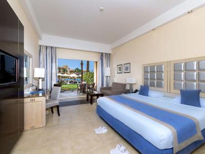Spacious hotel room with a king bed, blue accents, desk, and balcony view of a pool and garden.