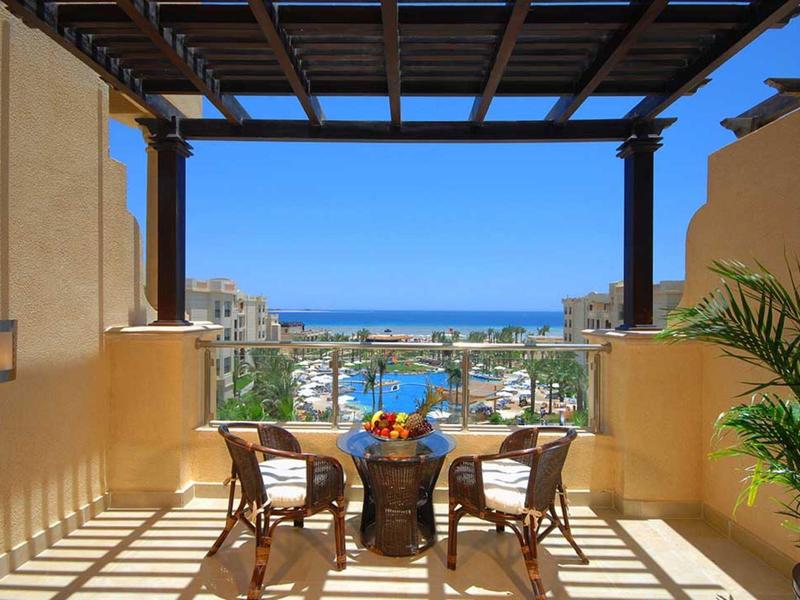 Balcony seating with table and chairs overlooking a coastal city and ocean view under a wooden pergola.