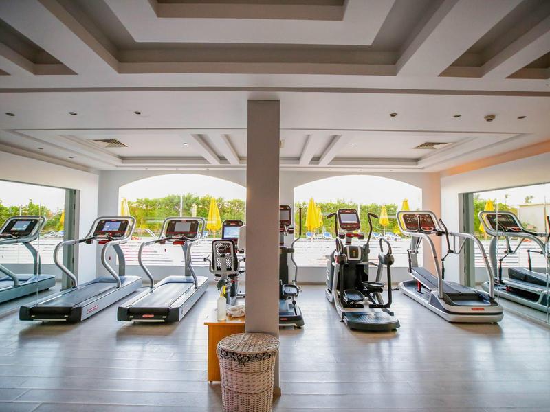 Modern gym with treadmills and elliptical machines, large windows showing greenery outside.