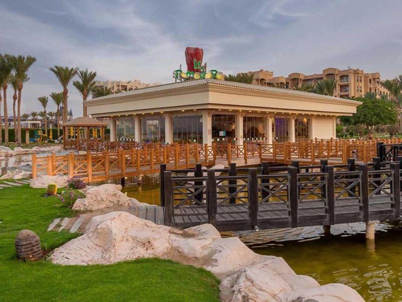 Outdoor restaurant by water with wooden bridge, green lawn, and palm trees under cloudy sky