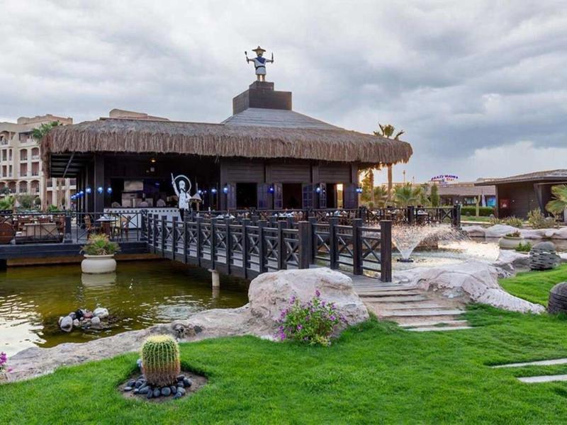 Resort with wooden bridge, pond, patio, and lush greenery under a cloudy sky.