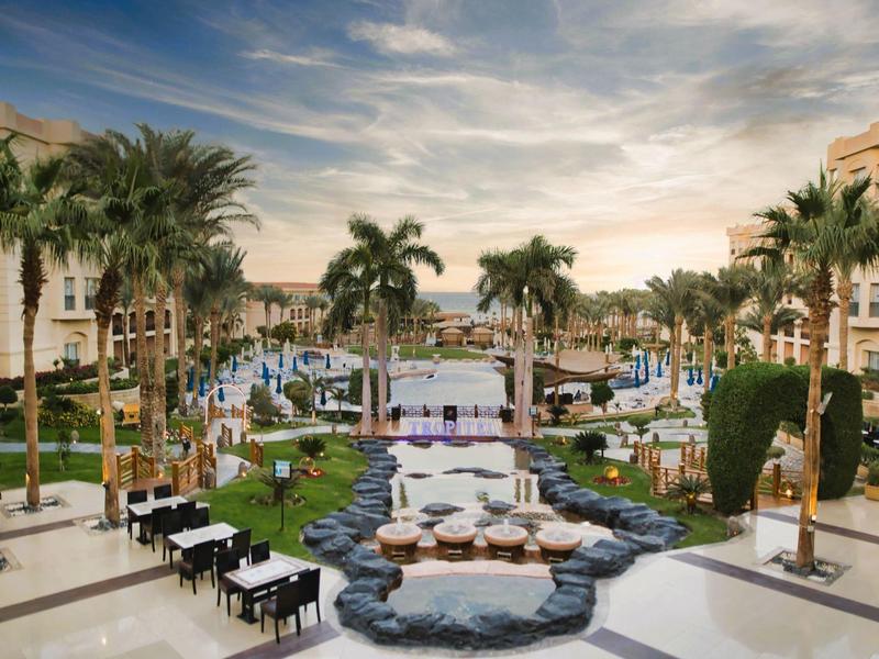 Resort pool area with palm trees, lounge chairs, and dining tables under a dramatic sky.