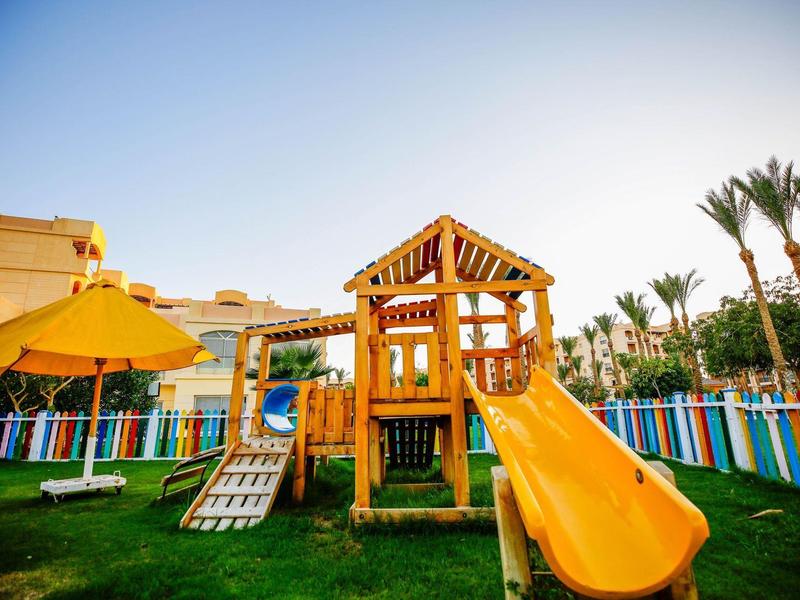 Wooden playground structure with yellow slide and umbrella in fenced grassy area under clear sky.
