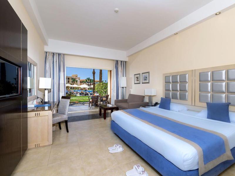 Spacious hotel room with a king bed, blue accents, desk, and balcony view of a pool and garden.