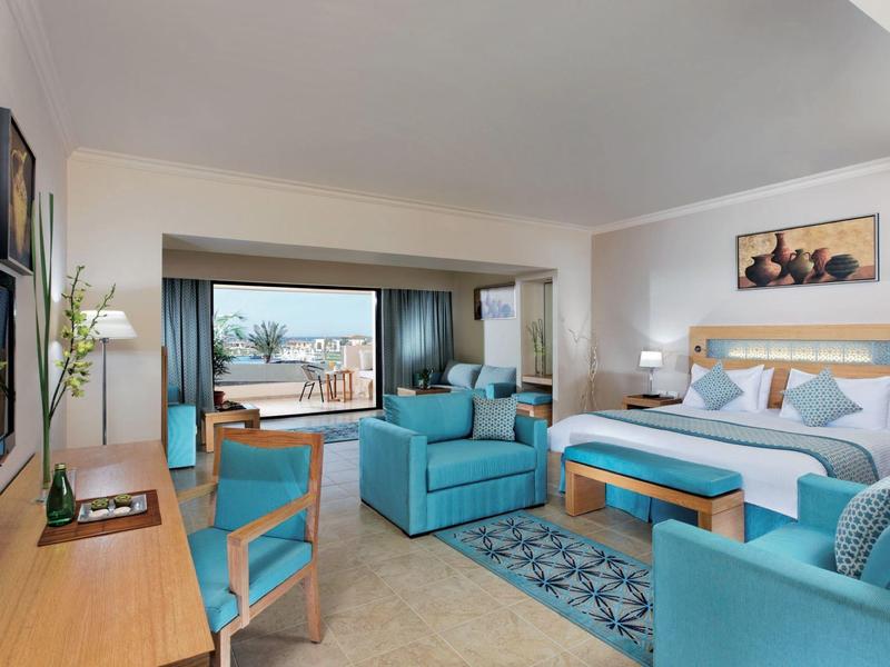 Helles Hotelzimmer mit blauem Sofa, Bett, Schreibtisch und großem Fenster mit Blick auf Terrasse.