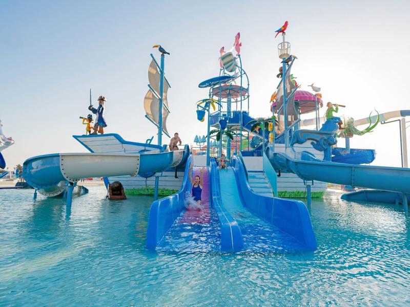 Wasserpark mit mehreren blauen Wasserrutschen und einem großen Spielgerüst im Pool unter blauem Himmel.