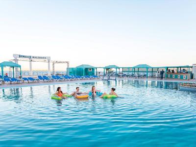 Dzieci pływają i bawią się w odkrytym basenie hotelowym z leżakami i parasolami w tle.