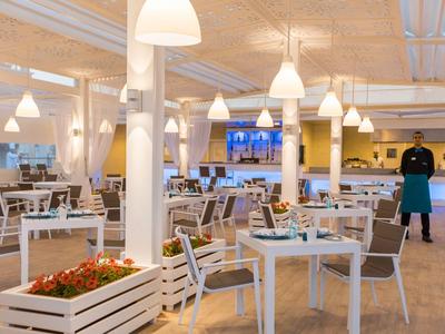 Nowoczesna restauracja z białymi stołami i krzesłami oraz dekoracyjnymi doniczkami.