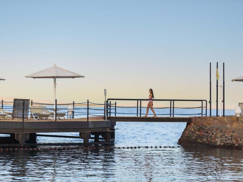 Eine Person geht auf einem Steg am Wasser mit Sonnenschirmen und klarem Himmel.