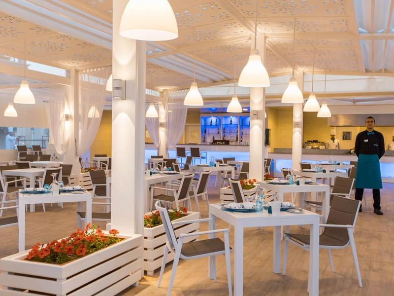 Helles Restaurant mit weißen Stühlen, gedeckten Tischen und einem Kellner im Hintergrund.