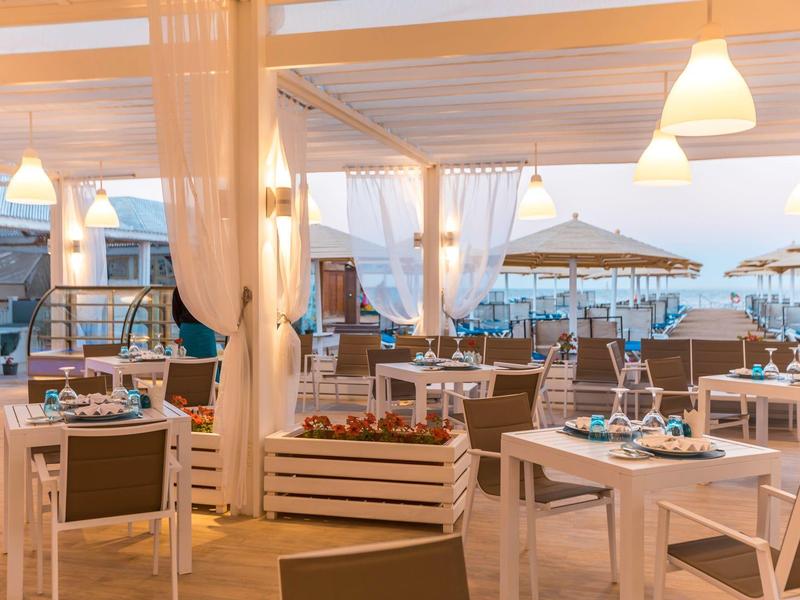 Elegantes Hotelrestaurant mit weißen Tischen, Stühlen und Blick auf das Meer bei Sonnenuntergang.