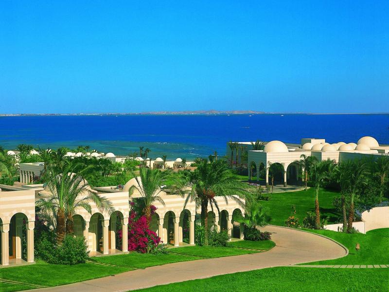 Resort with white buildings, palm trees, green lawns, and a sea view under a clear blue sky.