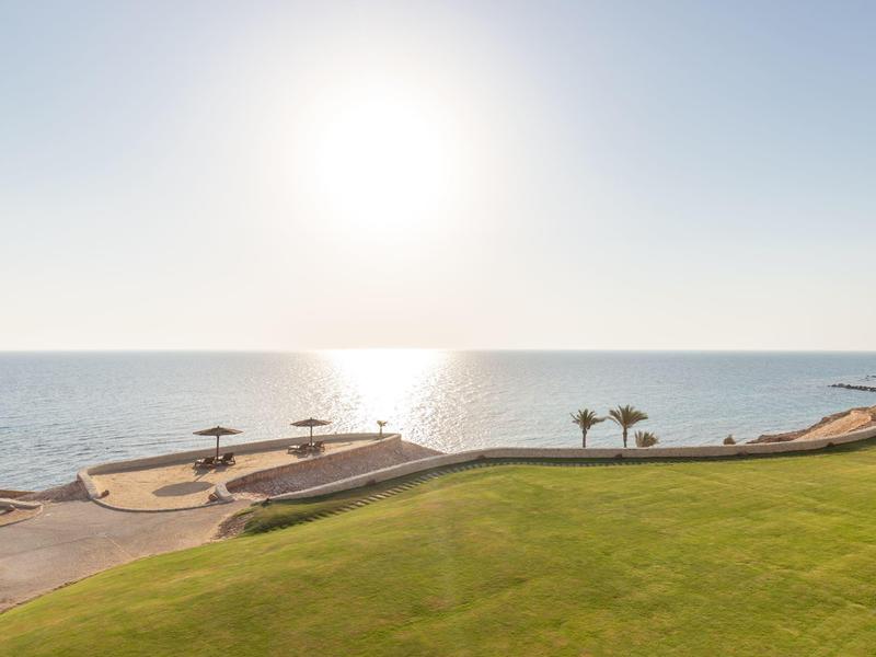 Sun shining over calm sea with grassy area and beach parasols in foreground.