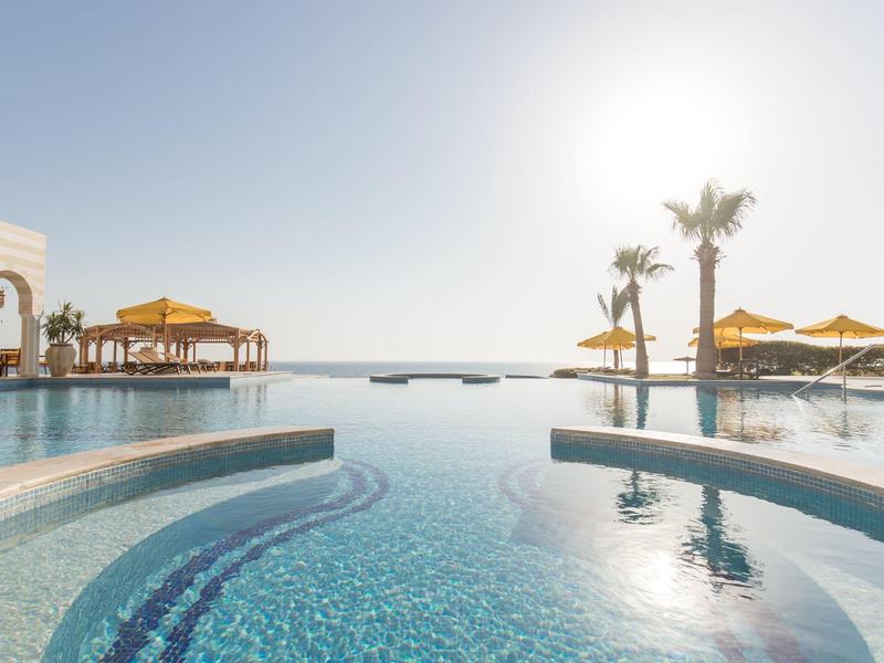 Infinity pool with umbrellas and palm trees under a clear sky near the sea.