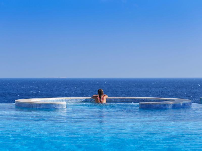 Person enjoying a circular infinity pool overlooking the ocean under clear blue sky.
